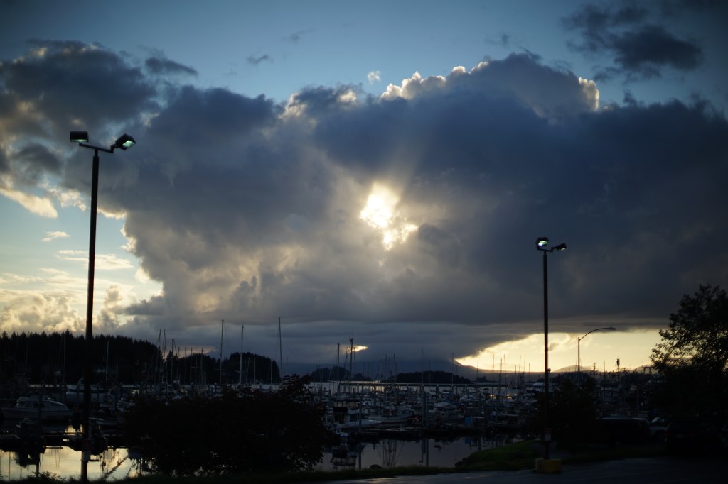 evening in a harbor parking lot, the ocean water in the harbor is still, the assembled masts of various boats reach calmly into the sky. and the sky, glowing silver and blue, a massive cloud hides the retreating sun behind it. in the cloud's center, an opening, the sun streams through. 