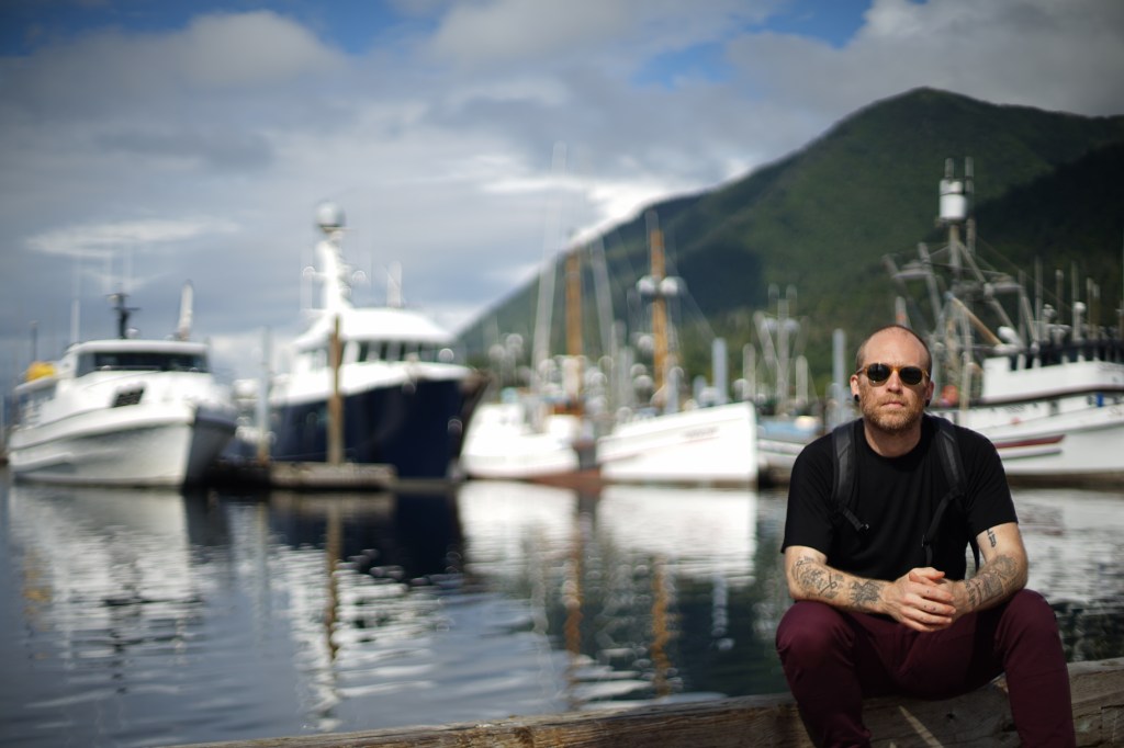 a man, thinning of hair but still looking moderately youthful, healthy even, sits in a black short sleeved shirt, maroon pants, and sunglasses, his arms wearing several black tattoos, on a dock. behind him, somewhat out of focus, large boats, water, a mostly-clouded sky, just a patch of blue, and a evergreen-tree covered hillside. some might call it a mountain.