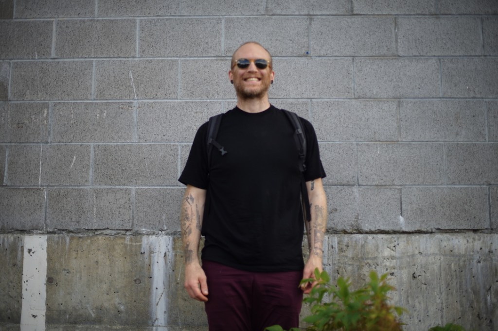 a man smiles broadly, dressed in a black shirt, maroon red pants, sunglasses, black tattoos, and auburn scruffy facial hair. behind him, a gray concrete brick wall. 