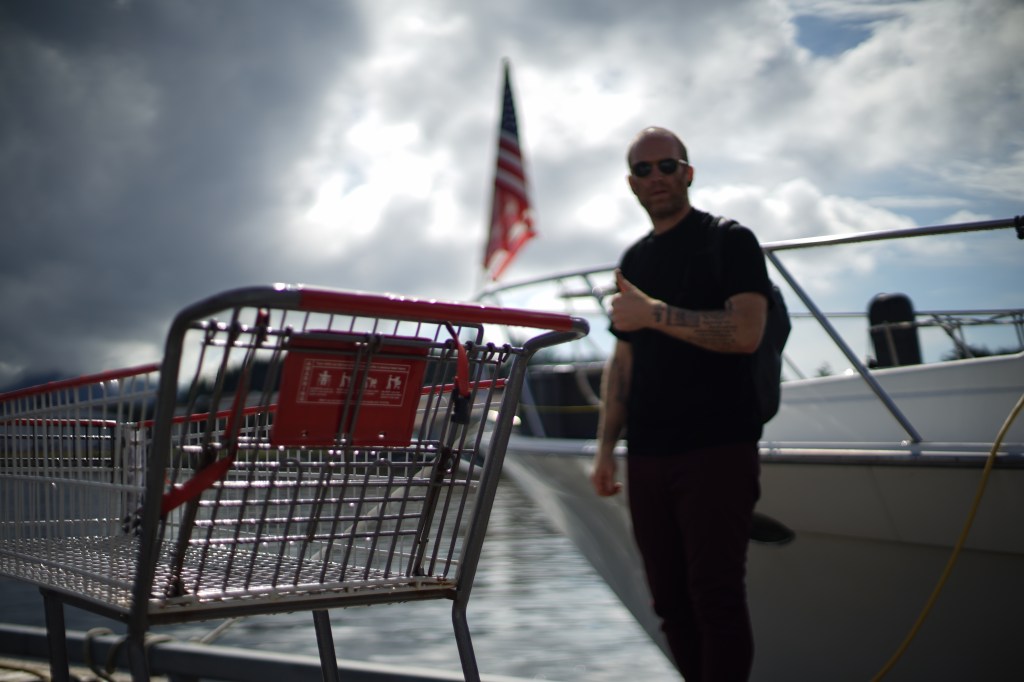 a man in sunglasses, long pants, and a short sleeved shirt, stands in front of the bow of a boat, giving a singly thumbs-up, an American flag limp and out of focus behind him. a shopping cart in the foreground, in focus and prominent. 