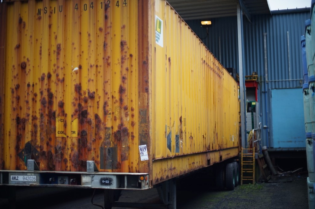 a yellow metal shipping container, backed up to a blue warehouse bay, rusts opulently like sunset on a rainforested island in the north pacific
