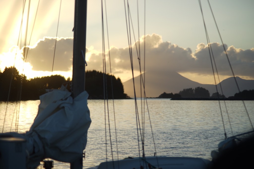 a sailboat, its mainsail furled, sits in the late evening setting sun. behind its chaos of lines and ropes and rigging, a gentle mountain rises in the distance, capped by billowing clouds. the sun shines brightly despite its near-setting. 