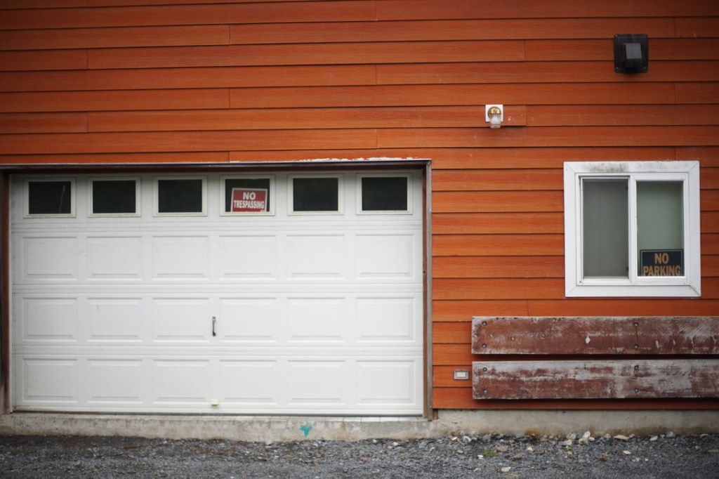 a rust-orange painted wooden building with a white garage door and white-trimmed window proclaims both "no trespassing" and "no parking' through signs in its windows.